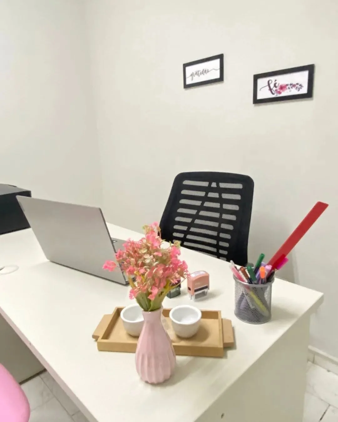 Mesa do consultório com notebook, vaso rosa com flores e os quadros gratidão e fé na parede