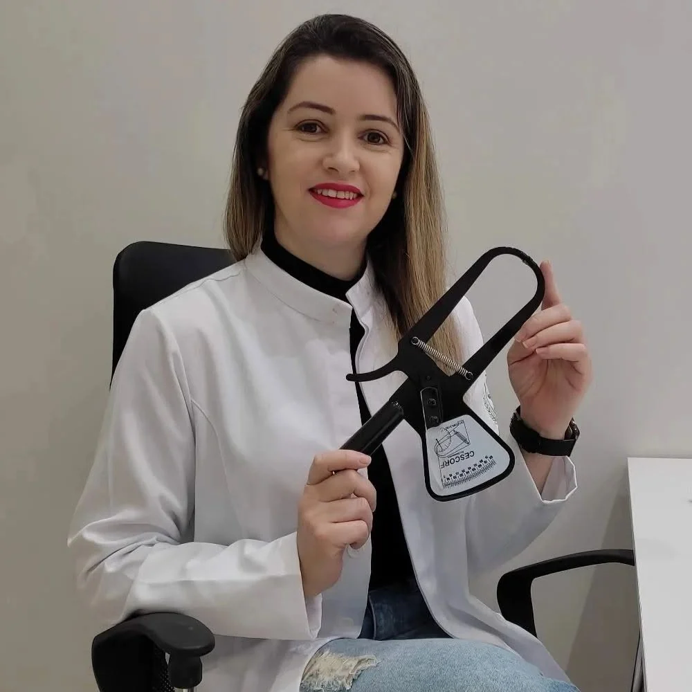 Gisleide de jaleco no consultório, mostrando o adipômetro usado na avaliação