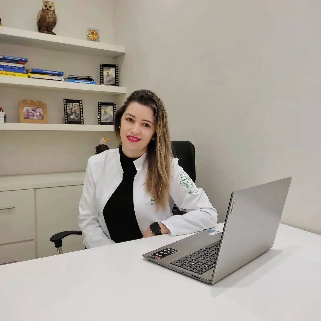 Gisleide sorrindo à mesa do consultório, de jaleco, com o notebook aberto